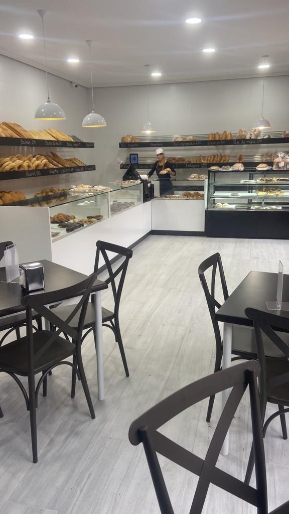 Un interior de panadería luminoso y moderno con un mostrador de cristal, estantes de madera con pan, mesas con sillas oscuras y un miembro del personal.