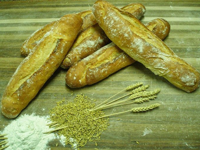 Varias baguettes doradas sobre una mesa de madera, junto a un montón de harina y tallos de trigo.