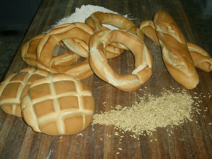 Una variedad de panes dorados, entre ellos rosquillas, panecillos y una baguette, reposan sobre una superficie de madera con harina esparcida.