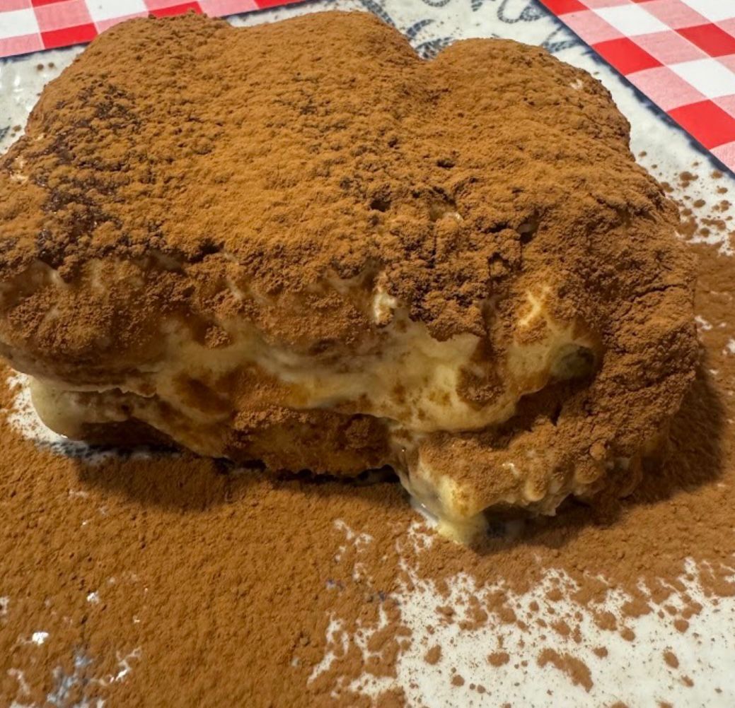 Postre de tiramisú, espolvoreado con cacao en polvo, sobre un plato con fondo de cuadros rojos.