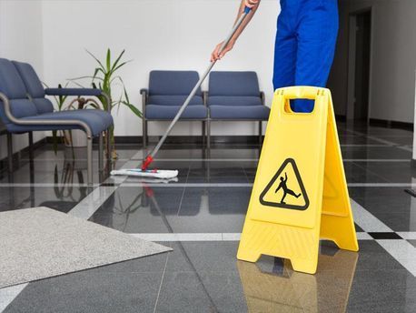 Persona trapeando un piso de baldosas brillantes al lado de un cartel amarillo de piso mojado en un área de espera.