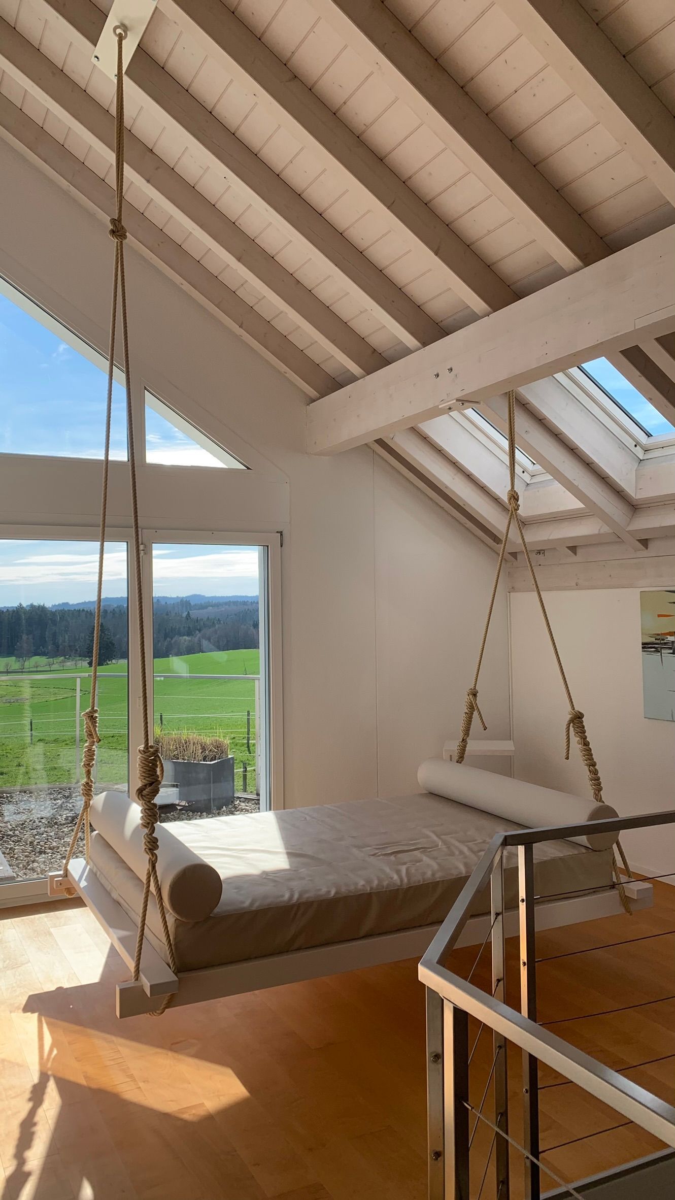 Ein an Seilen hängendes Bett in einem hellen, modernen Dachzimmer mit Oberlichtern und weitem Blick über grüne Felder.