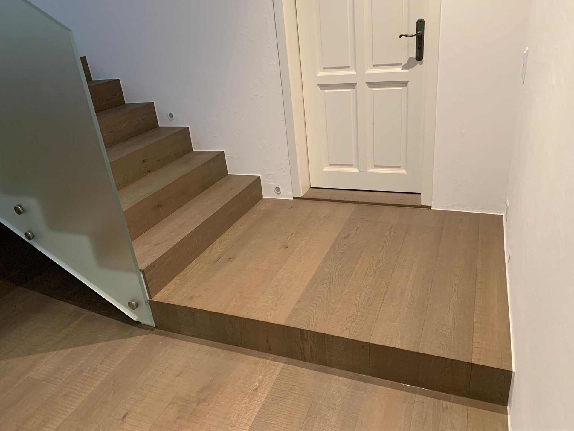 Moderne Holztreppe mit Milchglasgeländer, die zu einer weissen Tür in einem minimalistischen Flur führt.
