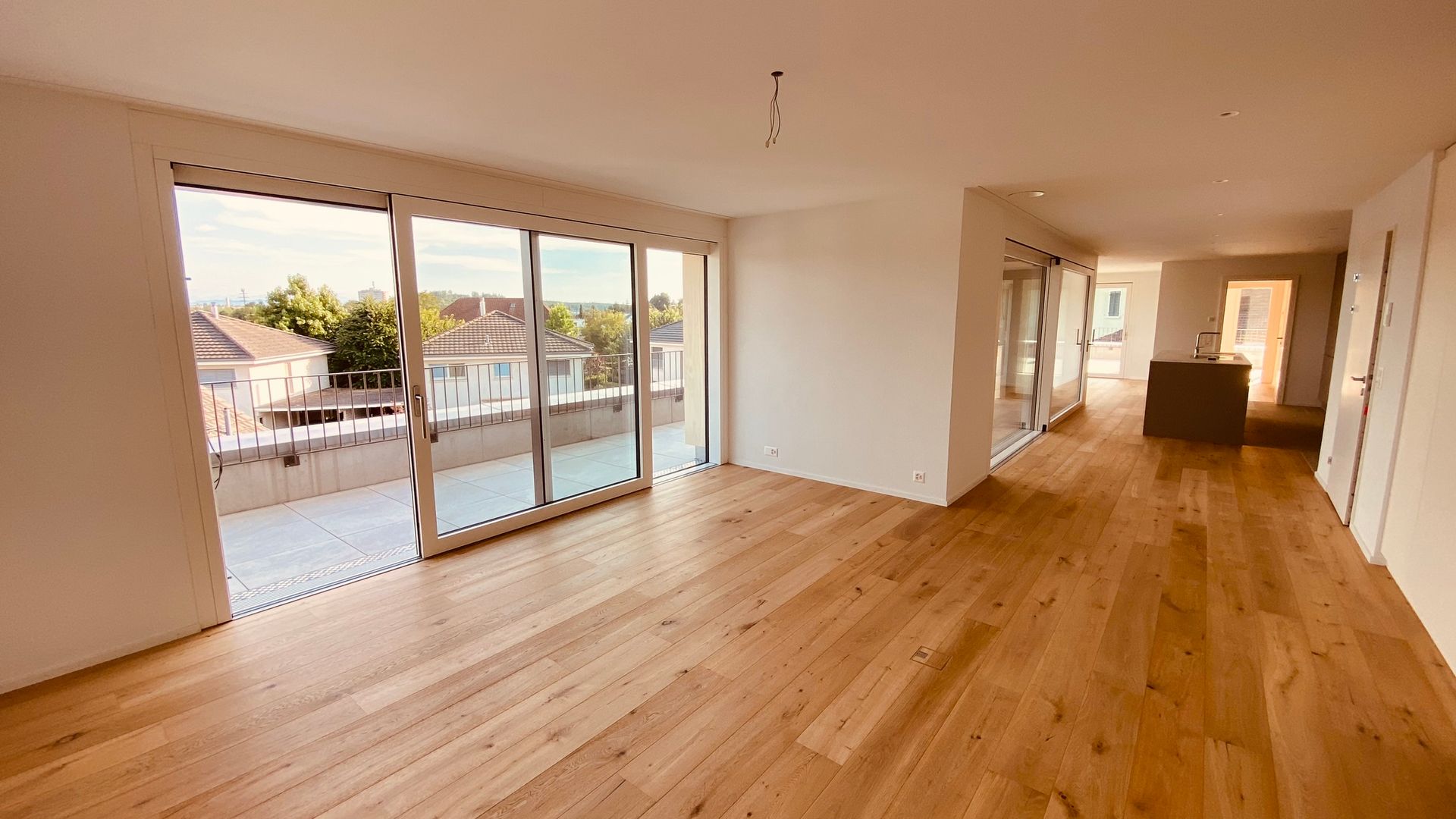 Ein leeres, modernes Apartmentzimmer mit hellen Holzböden, weissen Wänden und grossen, bodentiefen Glasschiebetüren.