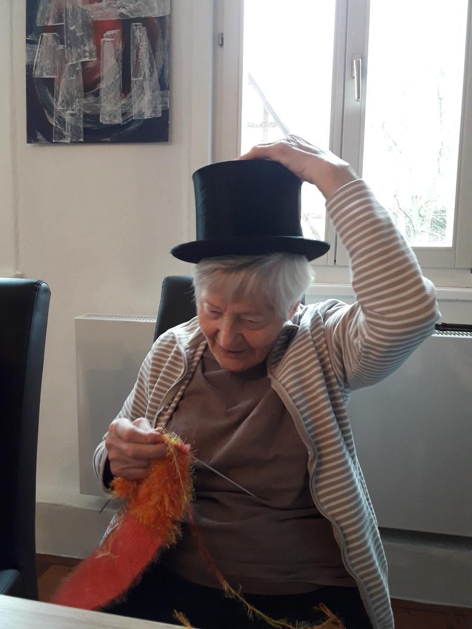 Seniorin strickt einen orangen Socken und trägt dabei einen schwarzen Zylinder auf dem Kopf