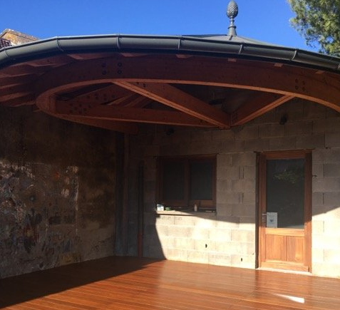 Une terrasse en bois semi-circulaire se trouve sous un toit incurvé assorti, prolongeant un bâtiment en pierre doté d'une porte en bois
