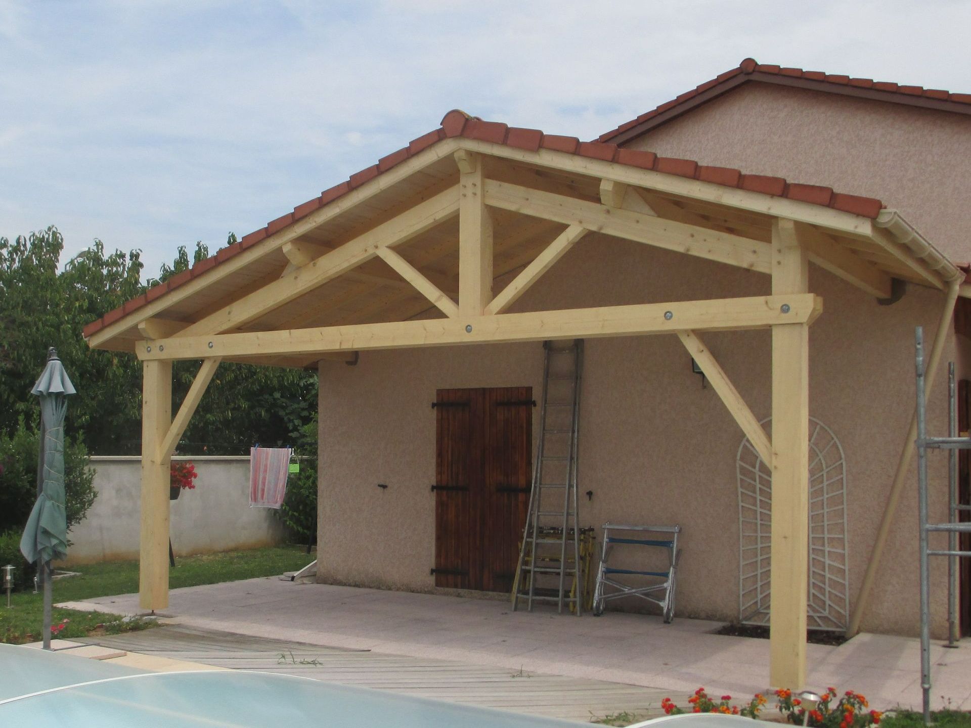 Une pergola en bois avec un toit en tuiles, adossée à une maison en stuc clair près d'une piscine