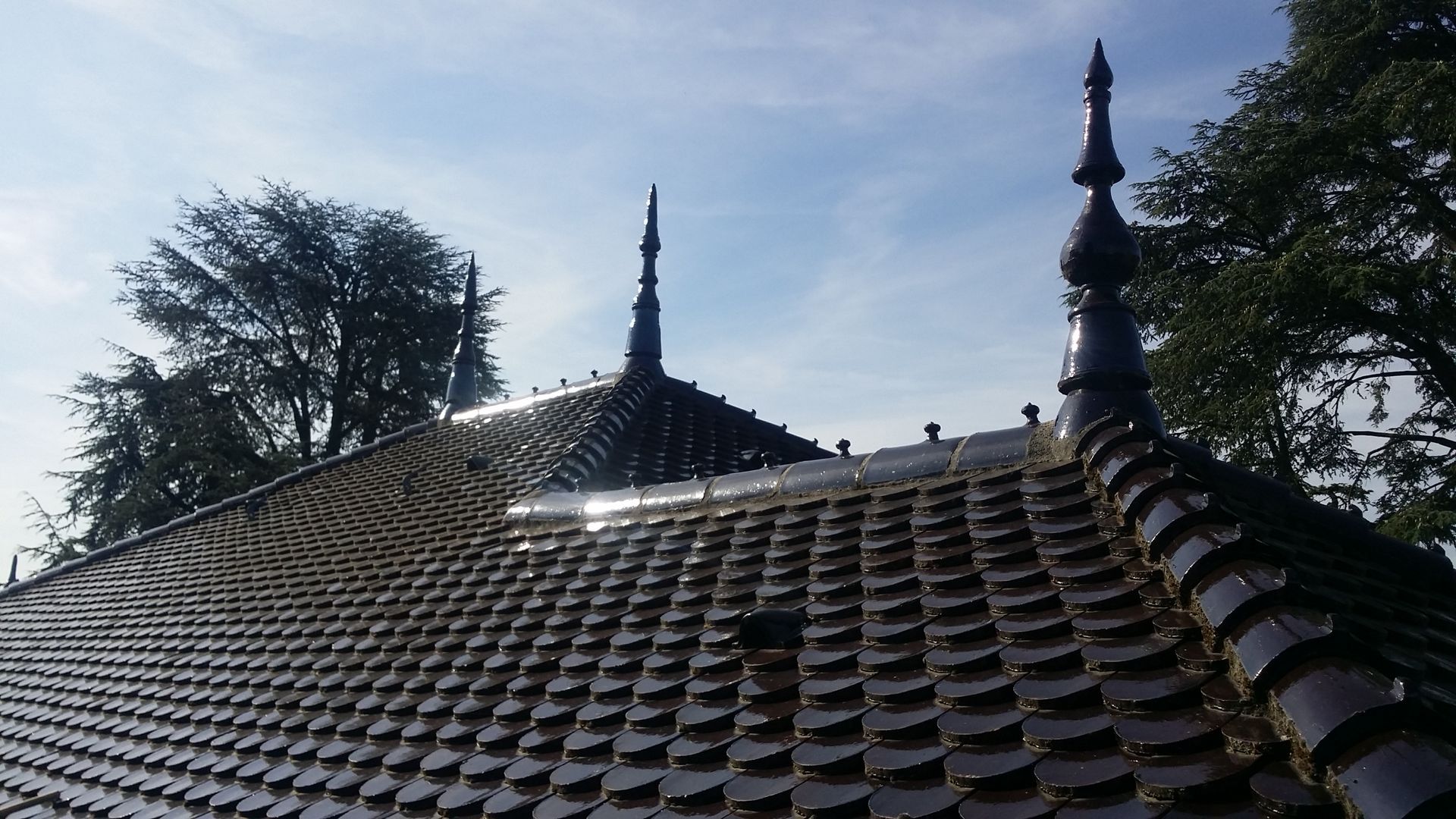 Vue en contre-plongée d'un toit de tuiles orné de trois pinacles en métal sombre, se détachant sur un ciel bleu et ensoleillé, des arbres en arrière plan