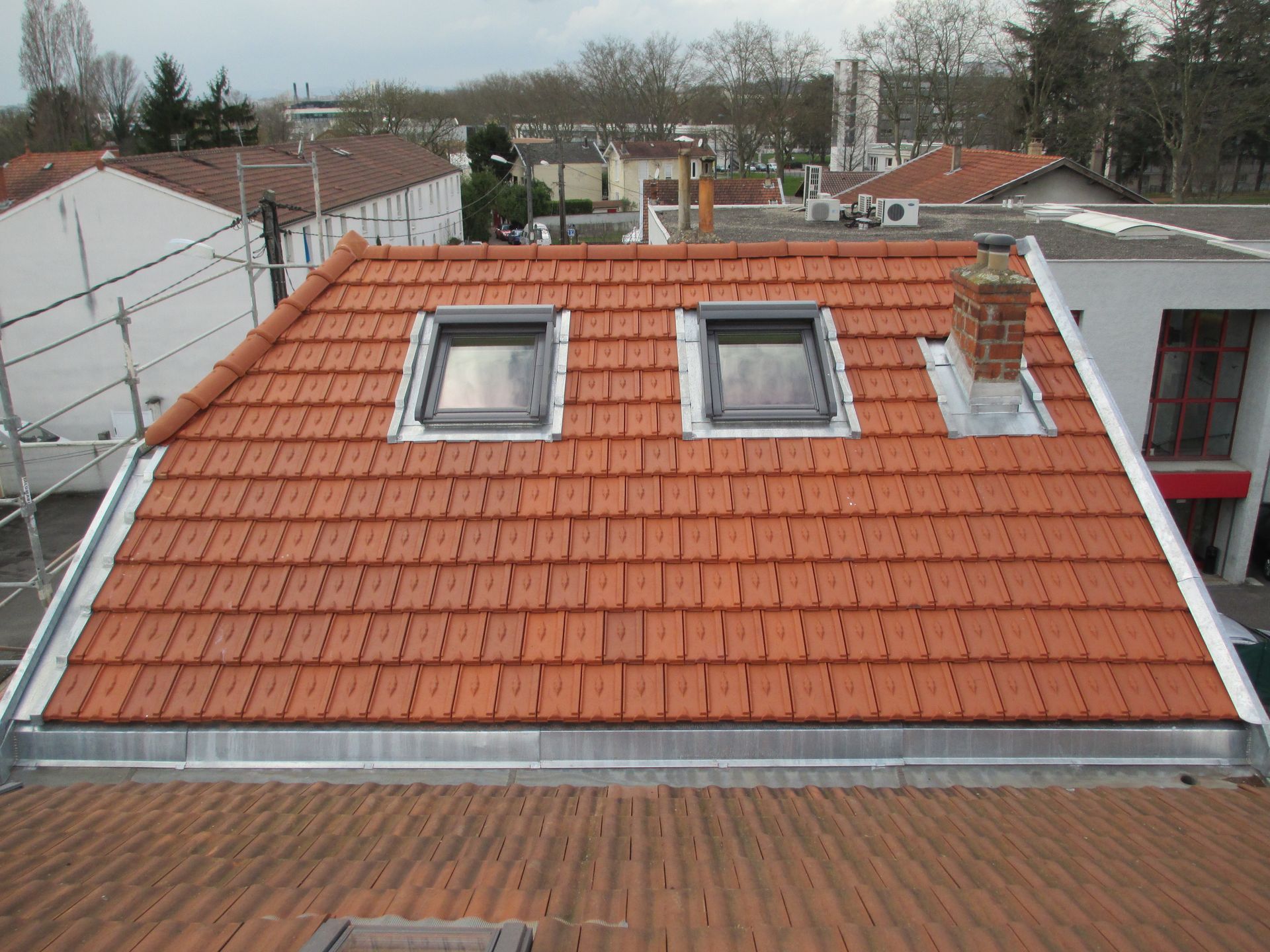 Vue du dessus d'un toit à deux pans recouvert de tuiles orange, deux puits de lumière et une cheminée en briques