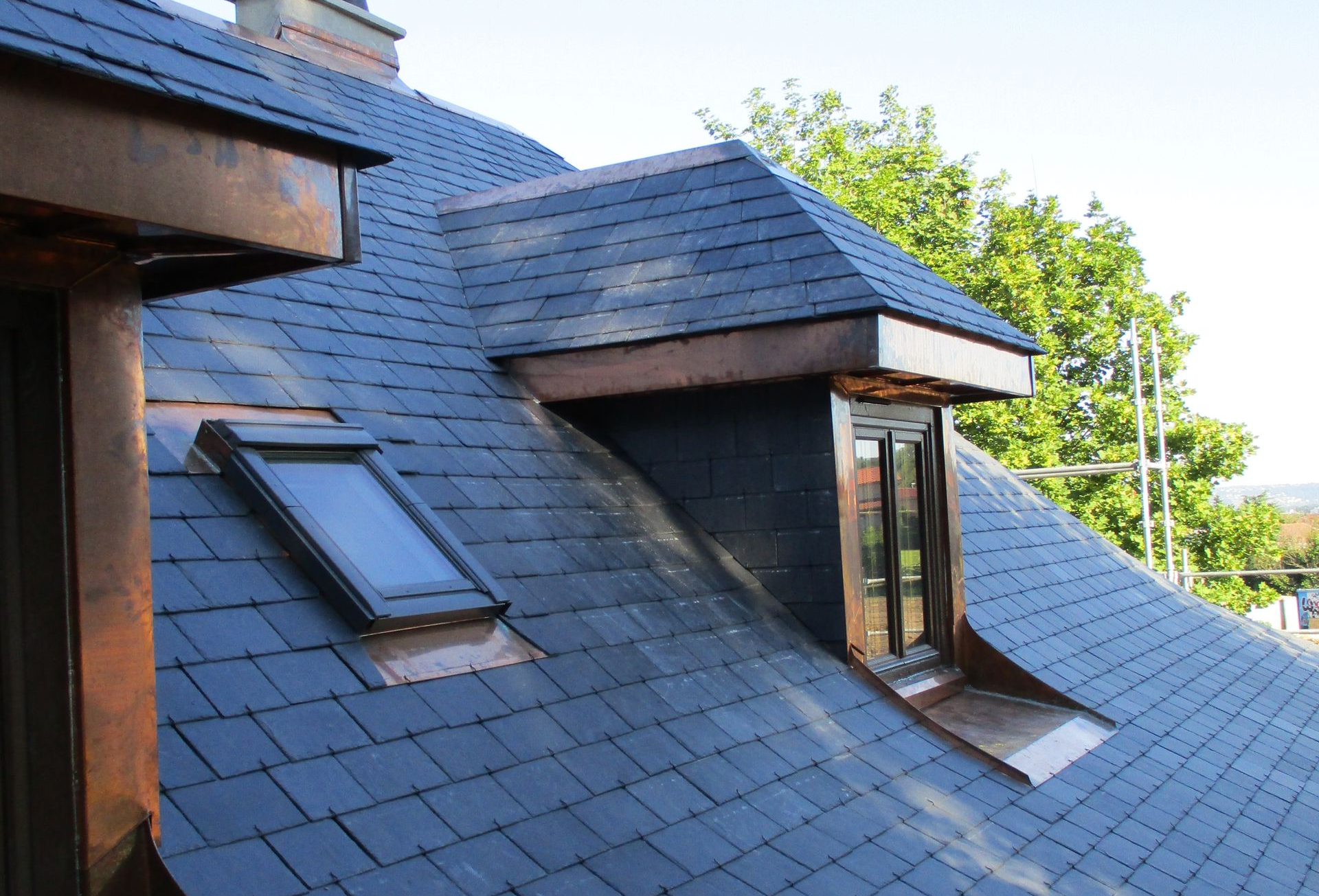 Un toit à deux pans en ardoise, percé d'un puits de lumière et d'une lucarne, se détachant sur un ciel bleu azur et une végétation luxuriante