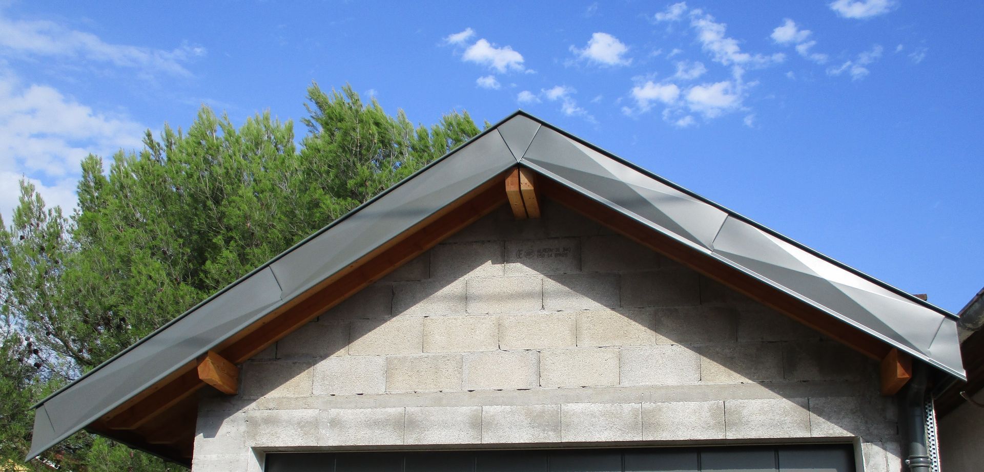 Le sommet d'un bâtiment en parpaings présente un toit en métal sombre, des chevrons en bois, des arbres verts se détachant sur un ciel bleu