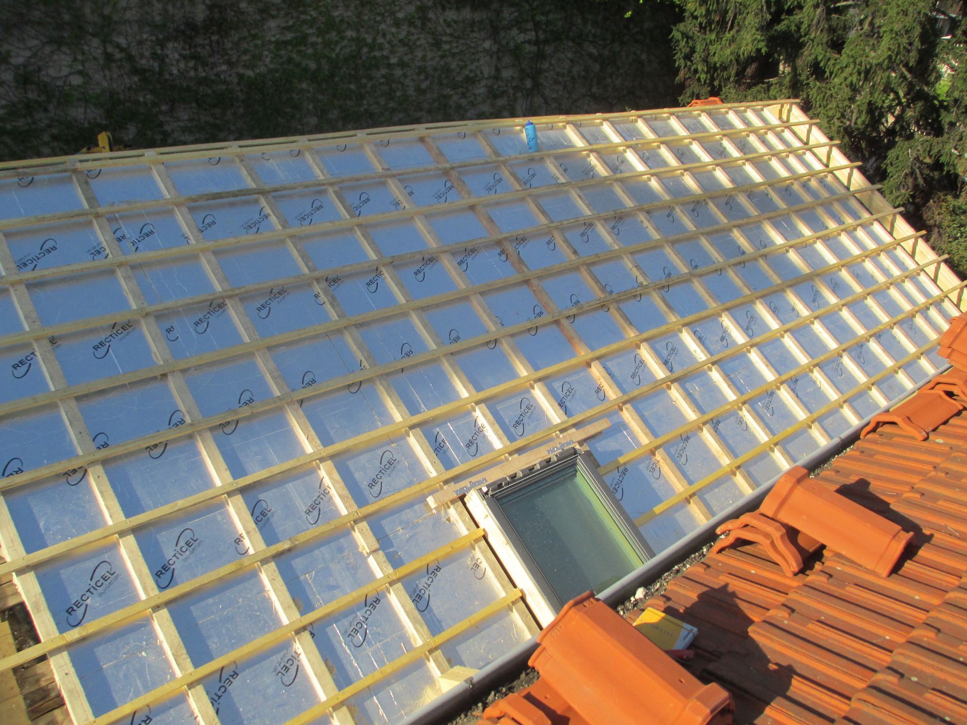 Un toit en pente en construction, avec des panneaux isolants et une charpente en bois, à côté de tuiles partiellement posées