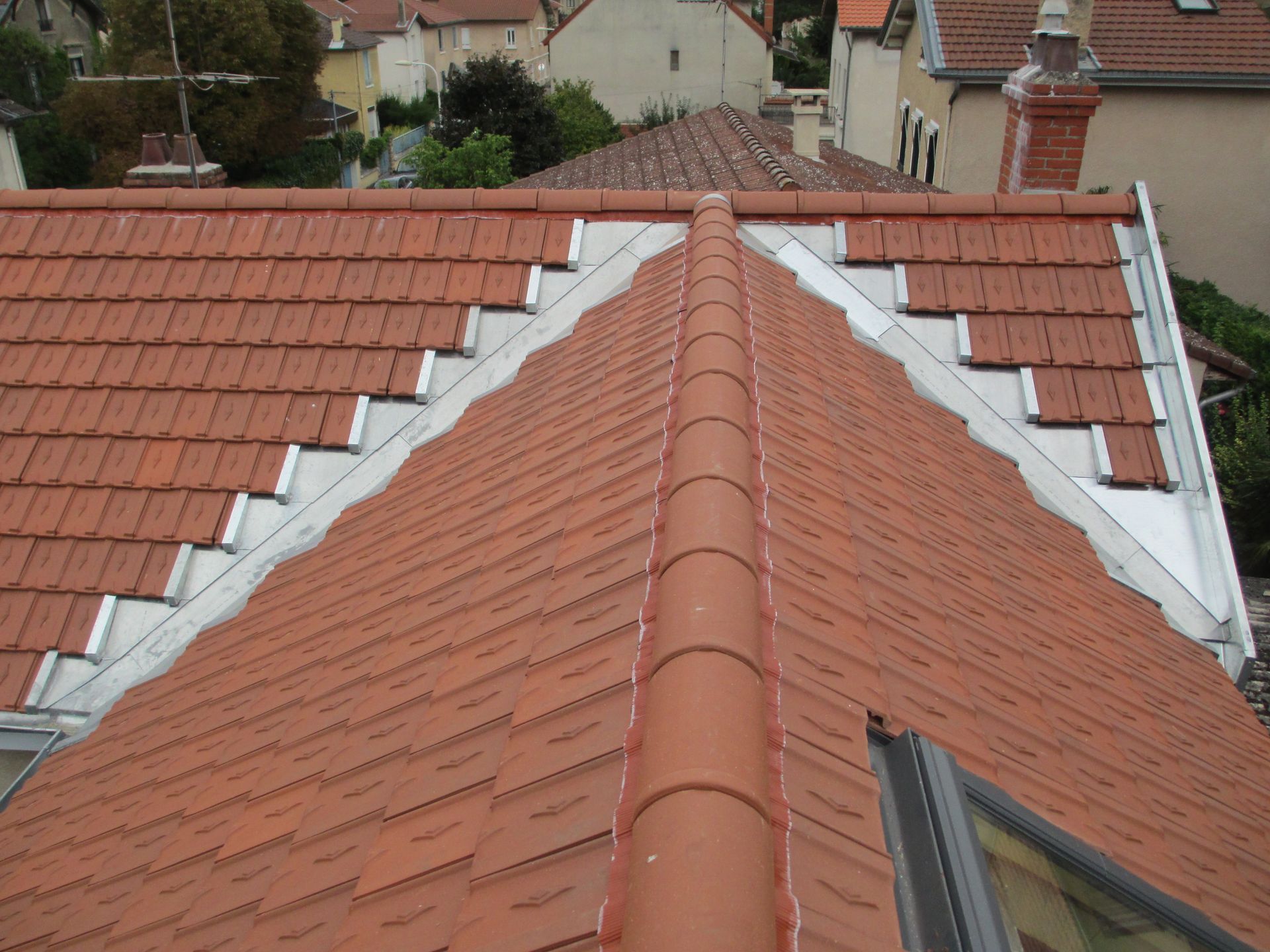 Vue en plongée d'un toit en tuiles rouges avec une arête centrale, des solins métalliques et un puits de lumière, se détachant sur un fond de maisons de banlieue