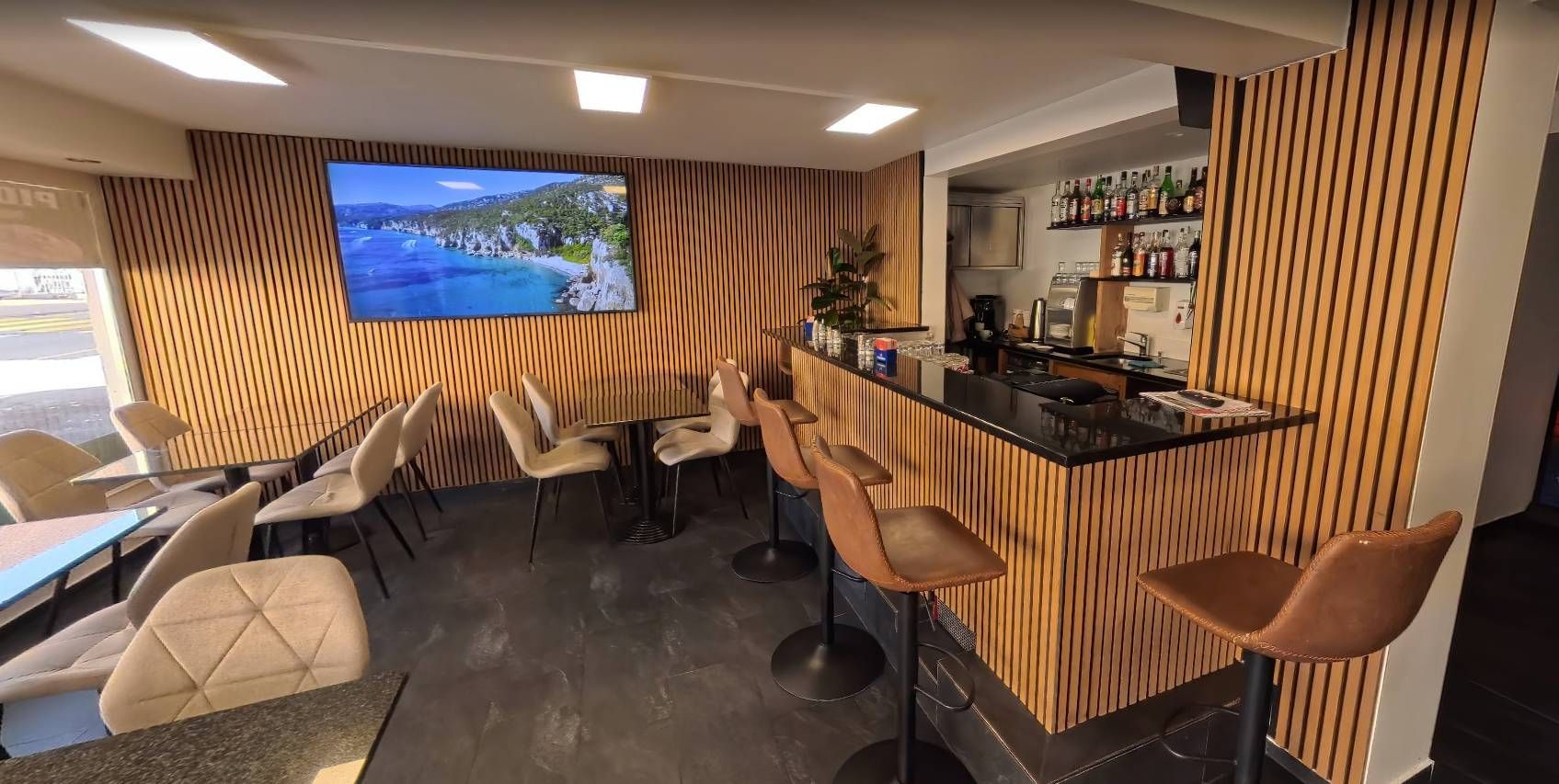 Innenansicht einer Bar mit schwarzer Theke, holzgetäfelten Wänden, braunen Barhockern, Tischen und einem Fernseher, auf dem eine Strandszene zu sehen ist.