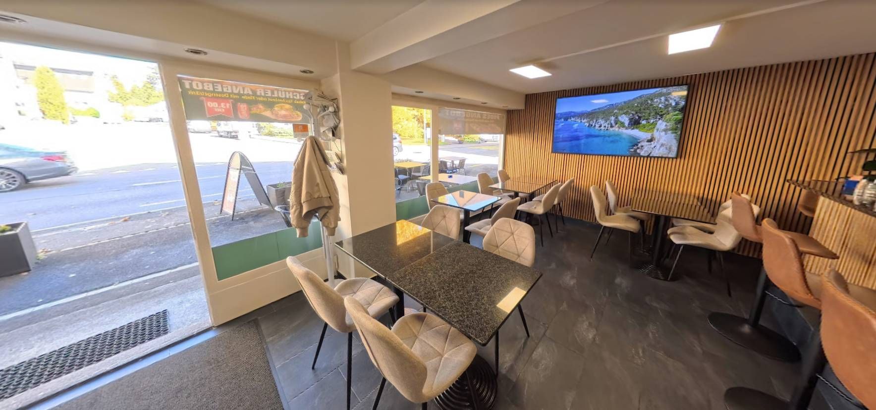 Innenansicht eines Cafés mit Tischen, Stühlen und einem grossen Fernseher mit Panoramablick. Die Fenster gehen auf eine Strasse hinaus.