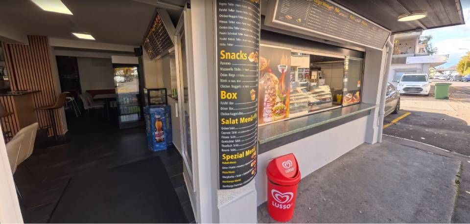 Aussenansicht eines kleinen Imbisses mit einer Speisekarte. Davor steht ein roter Mülleimer.