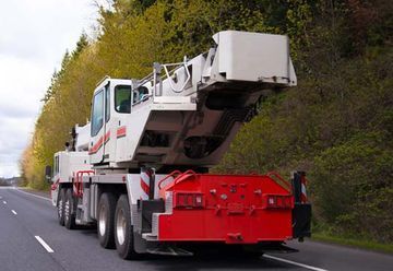 Un gran camión grúa blanco circula por una carretera junto a unos árboles, con un enganche delantero de color rojo.
