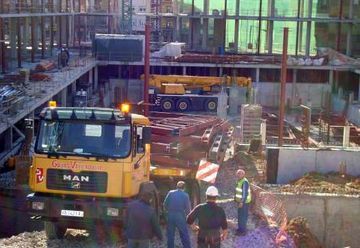Obra en construcción con camión MAN amarillo, trabajadores con cascos y una grúa junto a la estructura de un edificio.