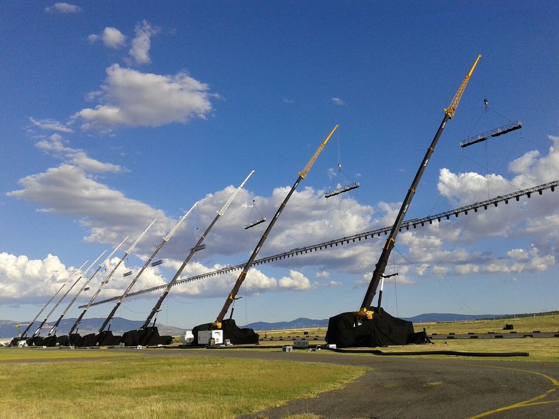 Una hilera de grúas izando una viga larga a través de un campo de hierba bajo un cielo azul con nubes.