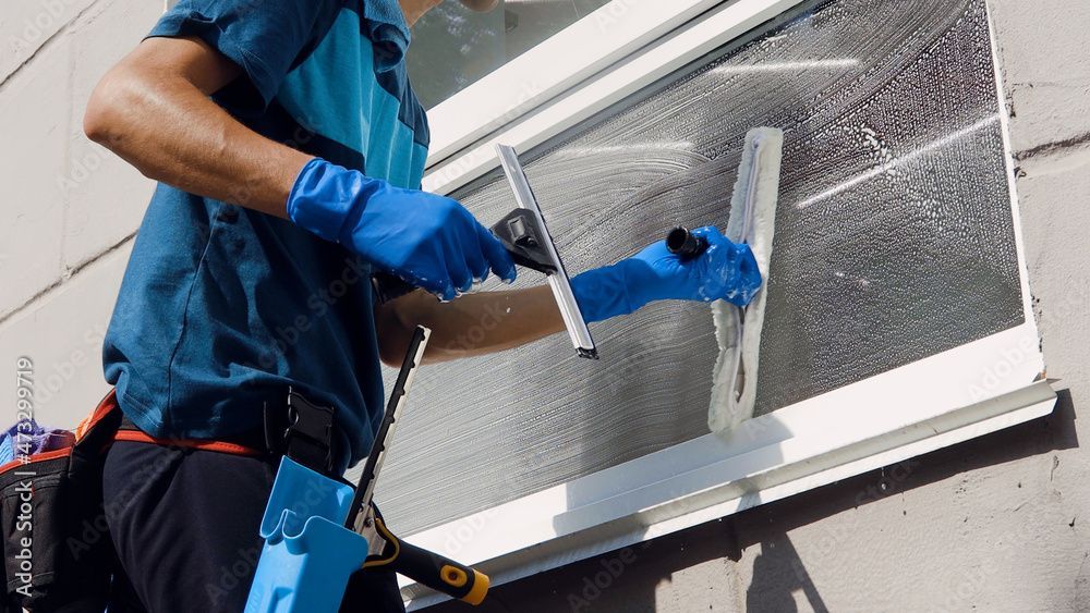 Eine Person mit blauen Handschuhen benutzt einen Abzieher, um ein Fenster vor einer hellen Gebäudefassade zu reinigen.