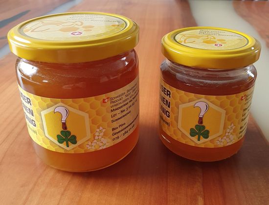 Zwei Gläser mit hellem Honig, ein grosses und ein kleines, mit gelben Etiketten und Deckeln, stehen auf einer Holzoberfläche.