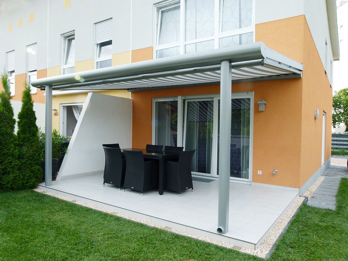 Eine Terrasse mit Tisch und Stühlen unter einem Vordach vor einem Haus.