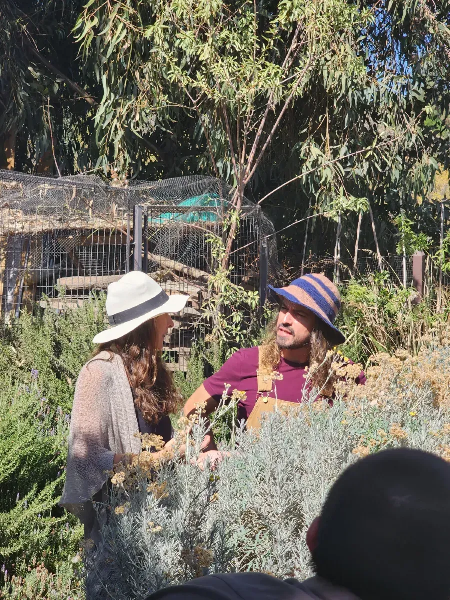 Una mujer y un hombre con sombreros conversan en un jardín, tras el follaje. El hombre lleva overol. Día soleado.