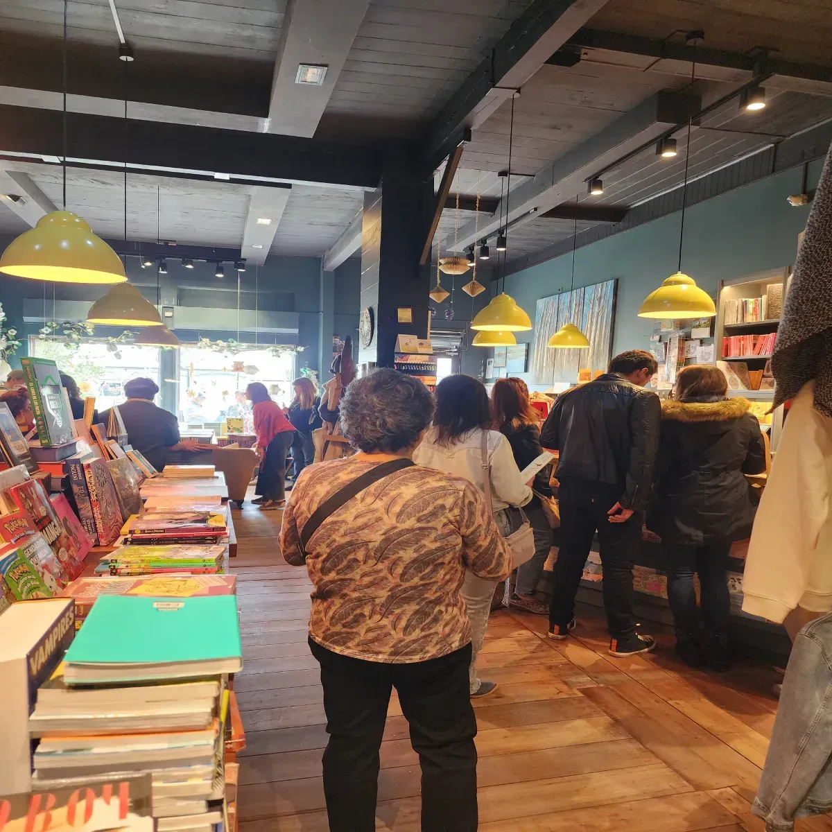 Dentro de una librería, la gente hojea estantes de libros bajo luces colgantes amarillas.