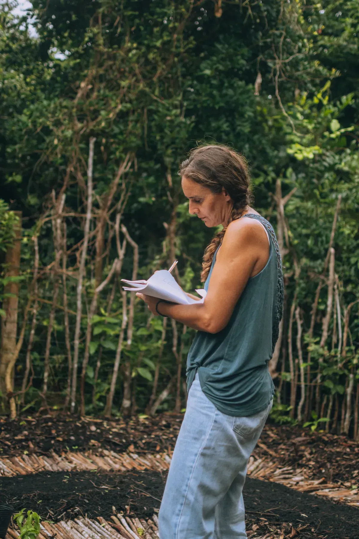 Mujer con camiseta verde y pantalones blancos, escribiendo en un cuaderno en un jardín.