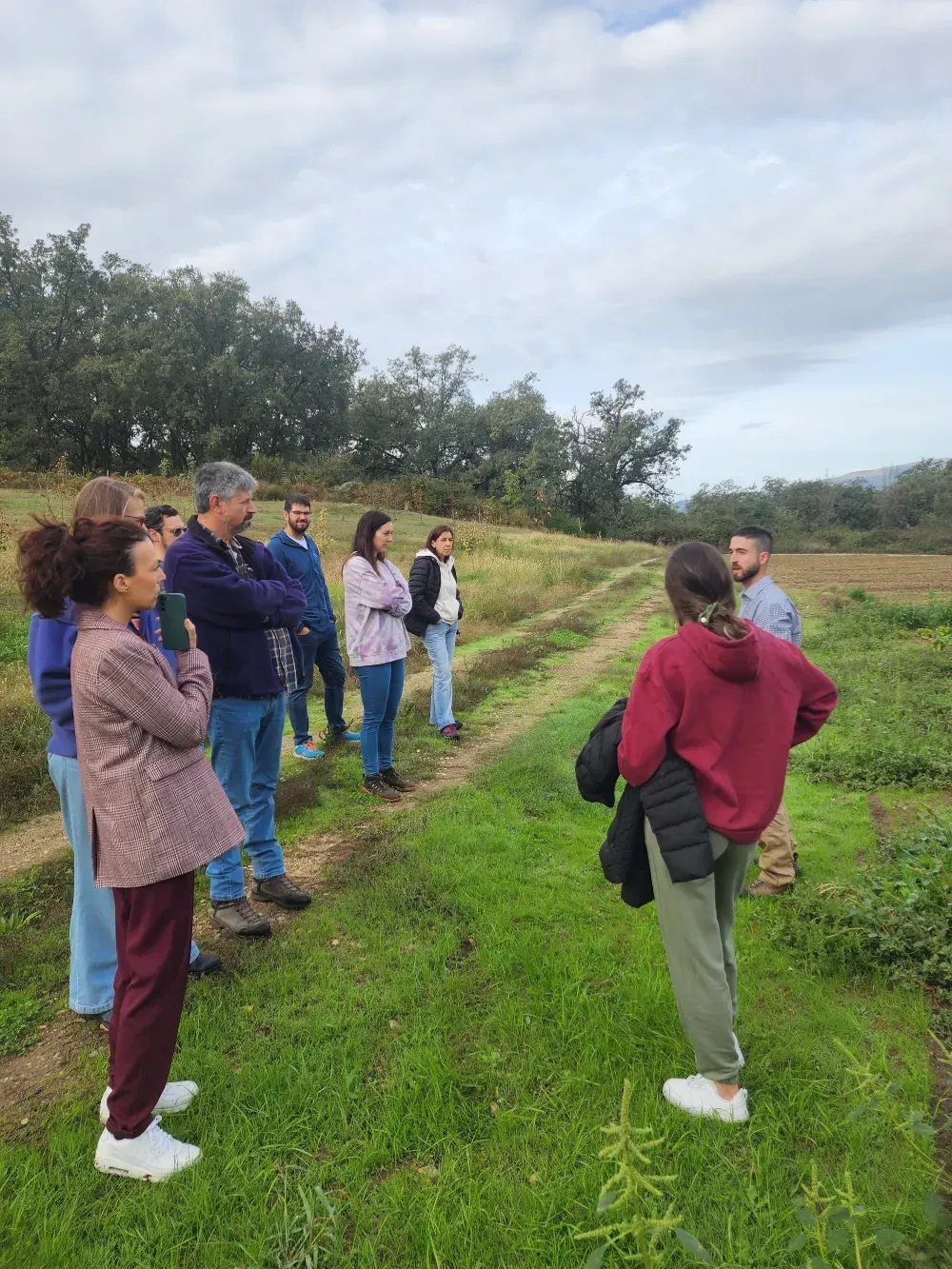 Un grupo de personas está de pie en un campo, escuchando a un hombre hablar. Cielo nublado, zona de hierba.