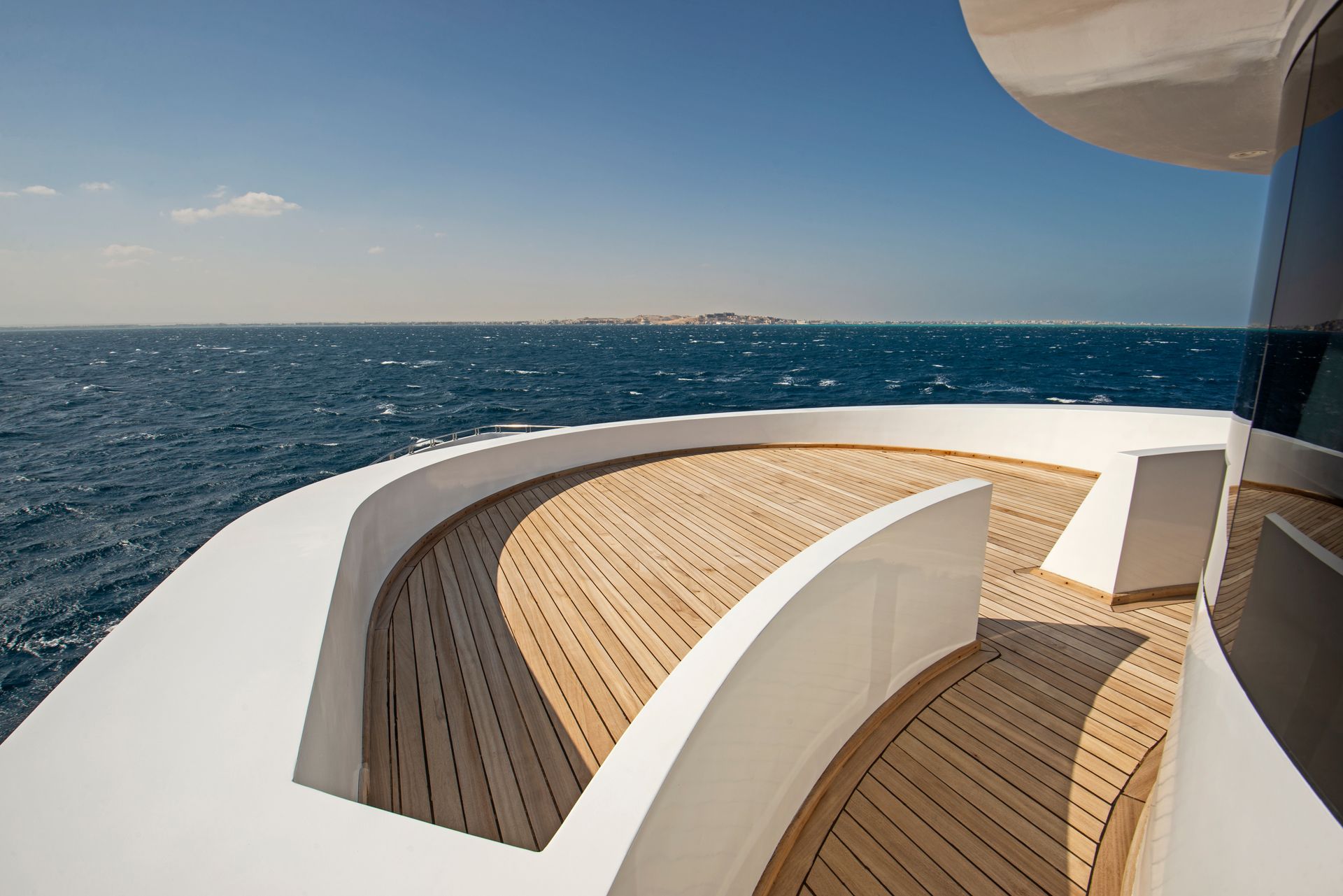 Luxuriöses Yachtdeck mit geschwungenen weissen Sitzbänken und Teakholzboden, mit Blick auf den offenen blauen Ozean unter klarem Himmel