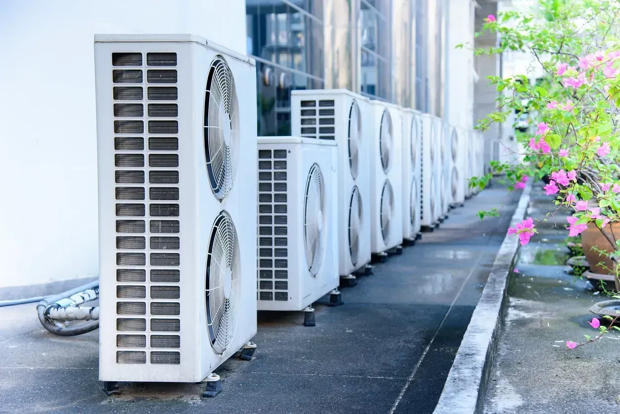 Una fila de unidades de aire acondicionado blancas alineadas contra un edificio, con una planta con flores rosas en el fondo.