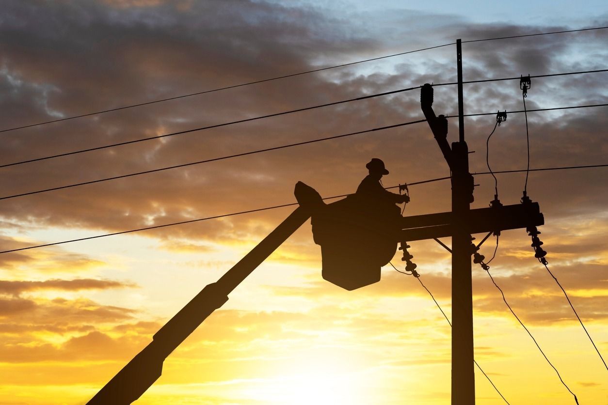 Silueta de un trabajador de servicios públicos en un camión cisterna trabajando en líneas eléctricas al atardecer.
