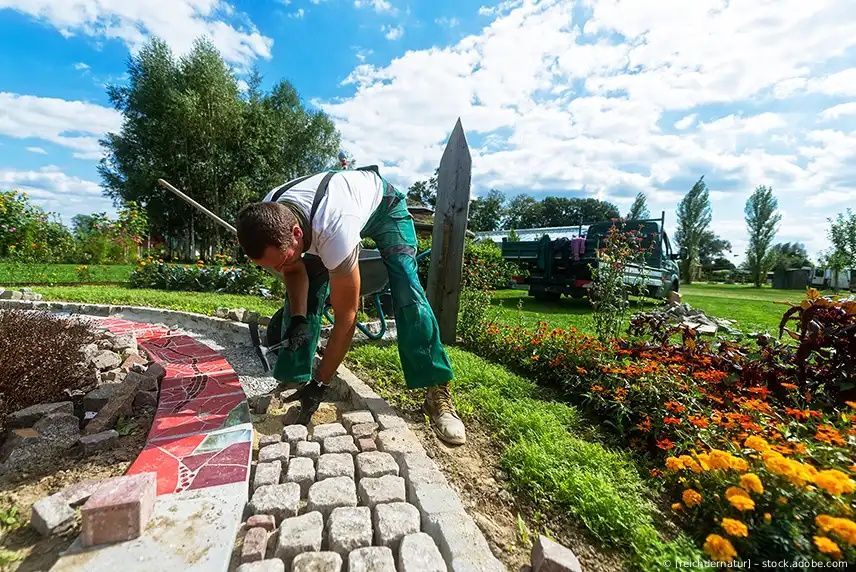 Ein Gärtner beugt sich über einen Kopfsteinpflasterweg und pflegt ein farbenfrohes Blumenbeet in einem sonnigen Park.
