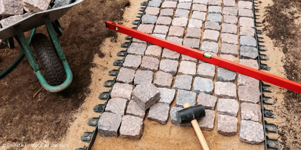 Ein Ziegelsteinweg wird mit Hilfe einer roten Wasserwaage und eines Gummihammers auf den Steinen verlegt.