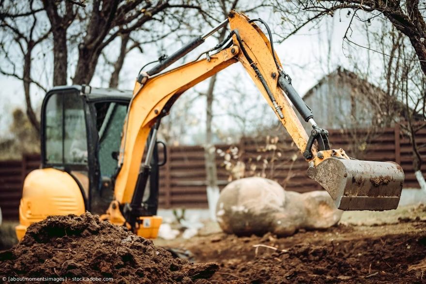 Ein gelber Bagger hebt Erde auf einer Baustelle aus; im Hintergrund sind kahle Bäume und ein Haus zu sehen.