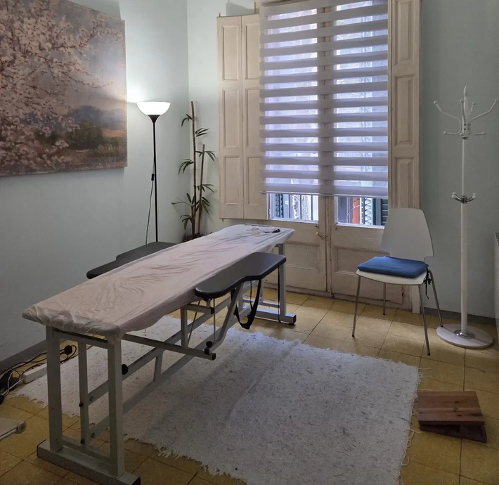 Sala de terapia de masajes con mesa de tratamiento, ventana, silla y perchero.