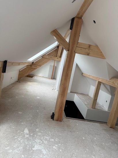 Une pièce mansardée inachevée avec des poutres de soutien en bois apparentes, des murs blancs inclinés