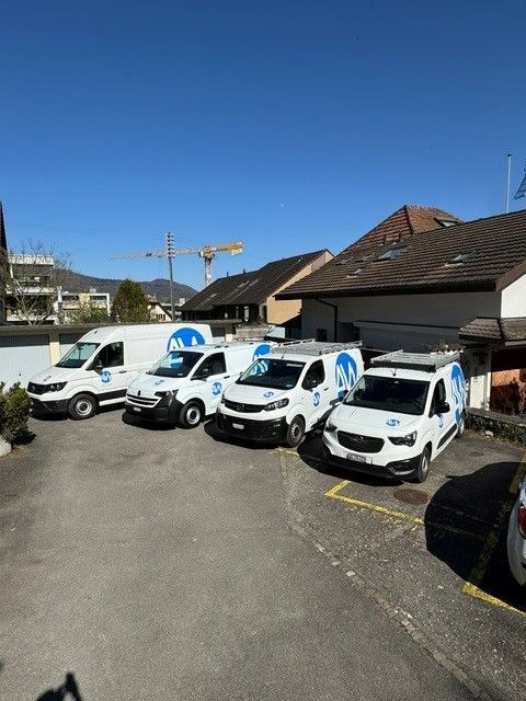 Draußen parken vier weiße Lieferwagen, jeder mit einem blauen Logo. Im Hintergrund sind Gebäude und Bäume zu sehen.
