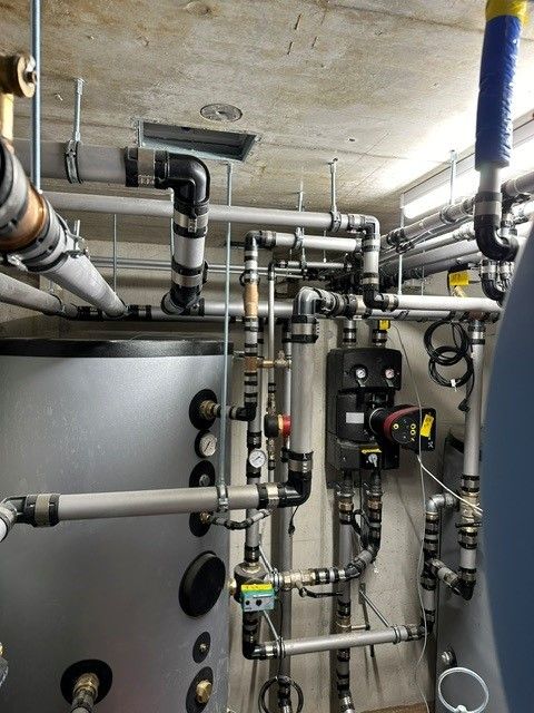 Komplexes System aus Rohren, Messgeräten und Pumpen in einer Industrieumgebung mit Metalltanks vor einer Betondecke.