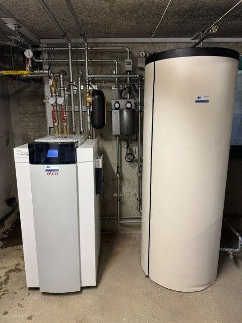 Heizsystem mit Heizkessel links, Warmwasserbereiter rechts sowie dazugehörigen Rohren und Bedienelementen an einer Betonwand.