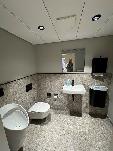 Öffentliche Toilette mit Urinal, Toilette, Waschbecken und Spiegel; Farbschema in Grau und Weiß.