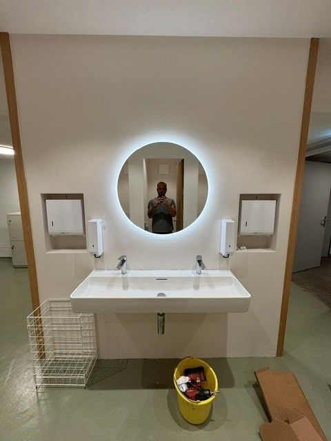 Badezimmer mit Doppelwaschbecken, rundem Spiegel mit Beleuchtung und dem Spiegelbild einer Person.