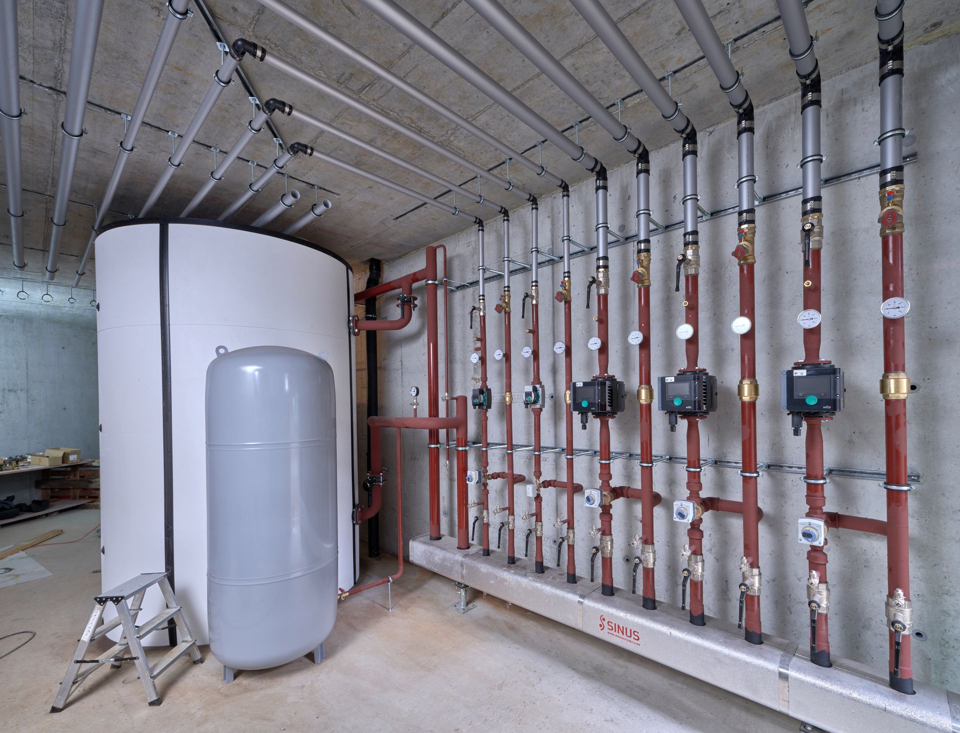 Komplexes System aus Rohren, Messgeräten und Pumpen in einer Industrieumgebung mit Metalltanks vor einer Betondecke.