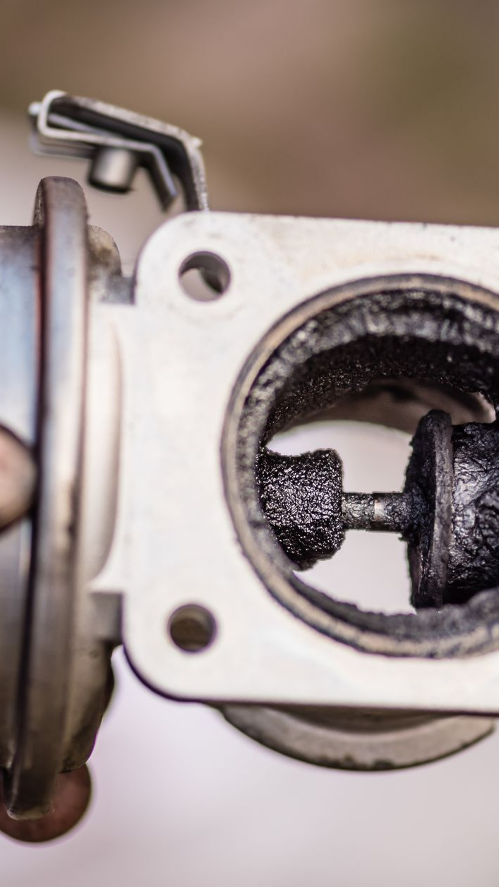 Gros plan sur une pièce automobile fortement recouverte de carbones, probablement une vanne EGR, tenue à la main sur un fond flou.