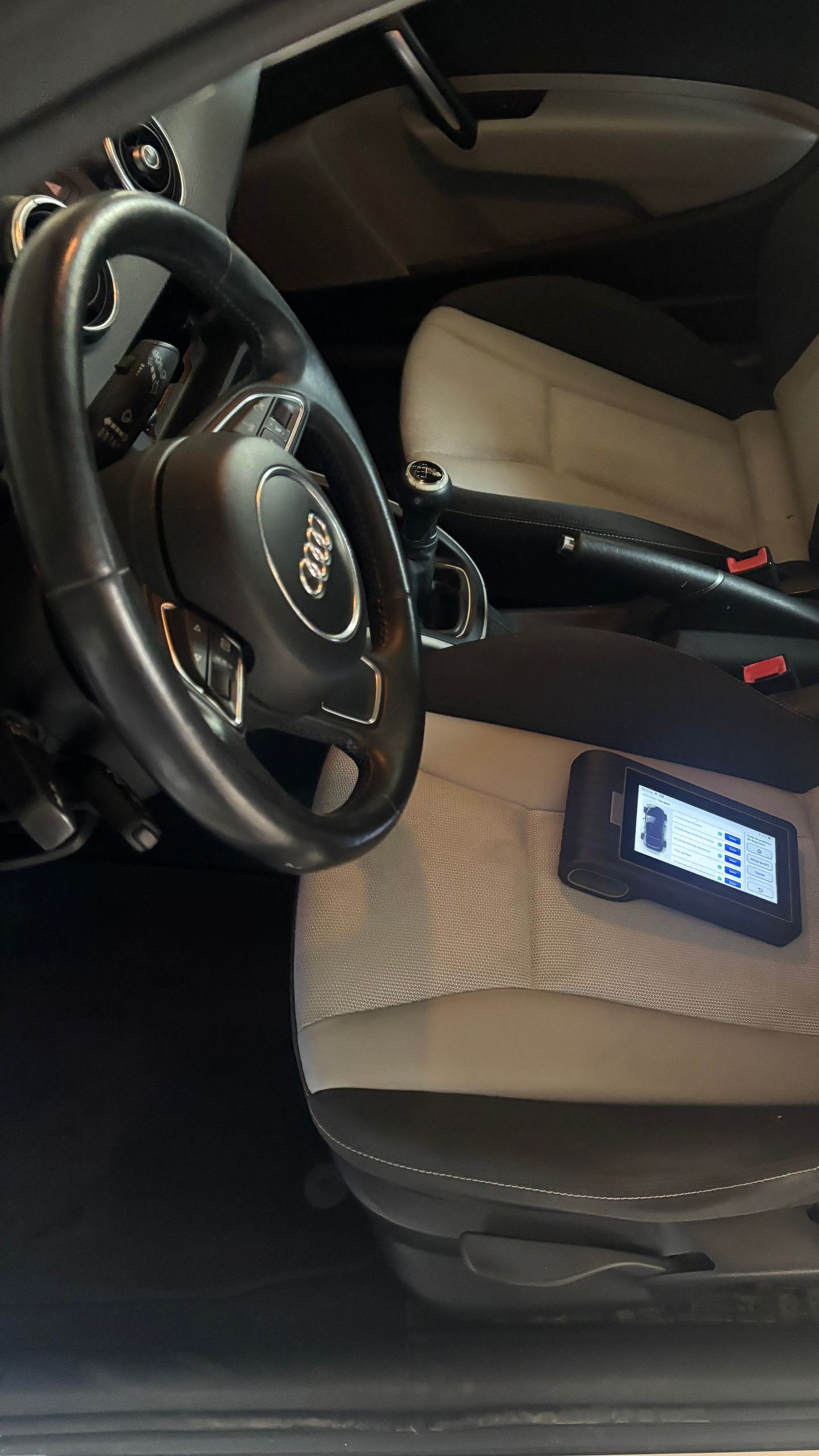Vue intérieure d'une voiture : volant, outil de diagnostic sur le siège. Sièges noir et beige, écran de l'outil allumé.
