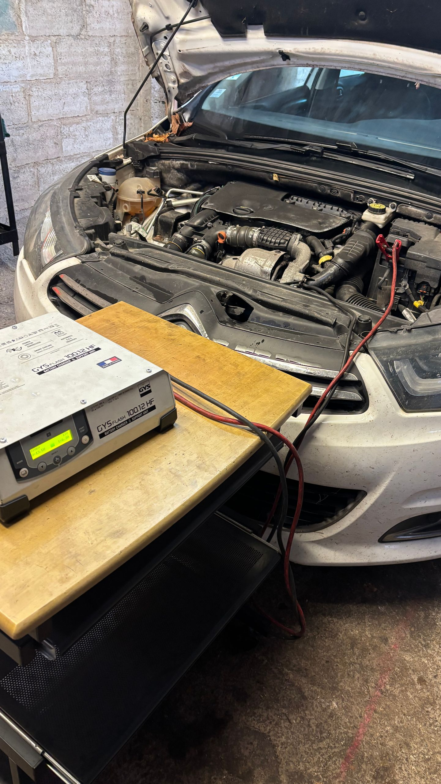 Le compartiment moteur de la voiture est ouvert, avec un chargeur connecté à la batterie, sur une planche en bois.