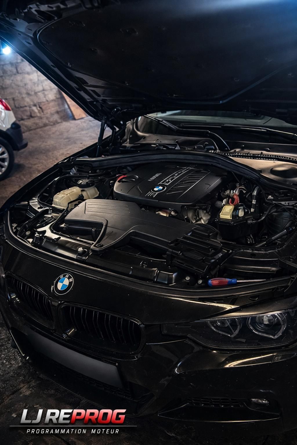 Une BMW noire, capot ouvert, est garée dans un garage, laissant apparaître le moteur. La voiture est immatriculée en France.