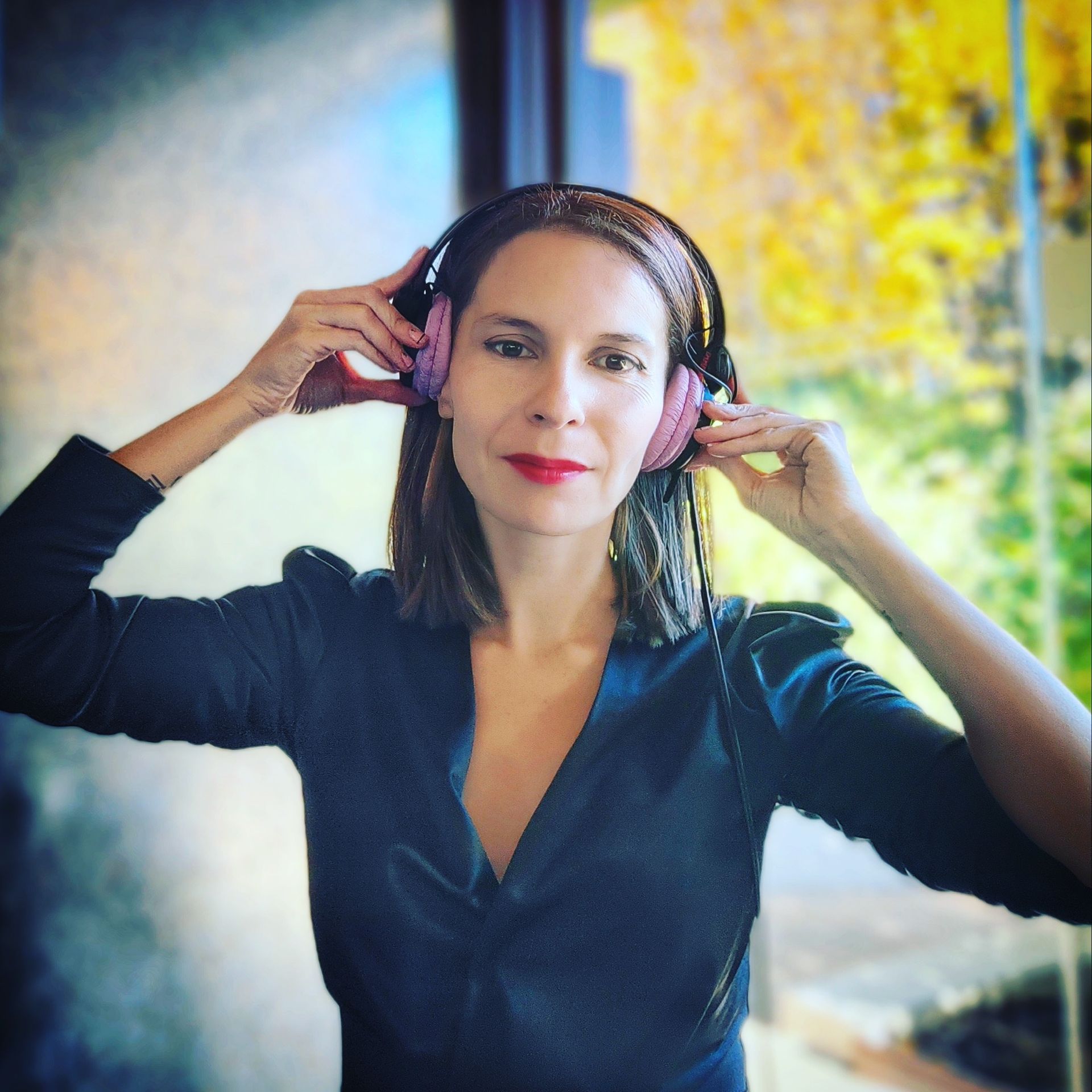 Mujer con auriculares rosas, sosteniéndolos en alto. Lleva camiseta negra y lápiz labial rojo. Fondo: ventana y árboles.