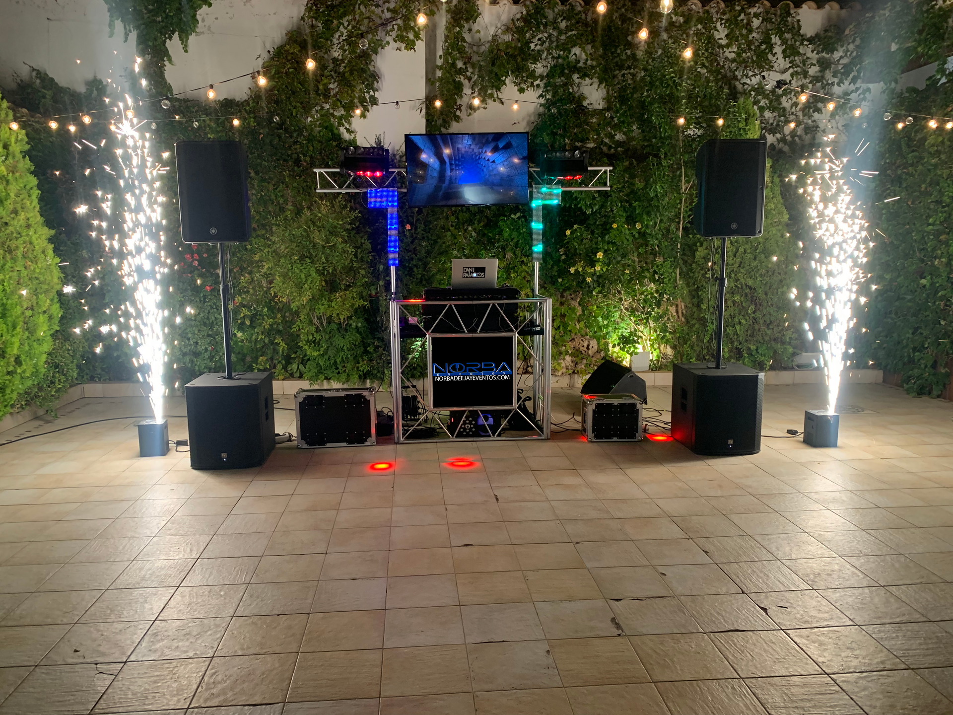 Configuración de DJ con altavoces, pantalla y pirotecnia al aire libre en un patio.